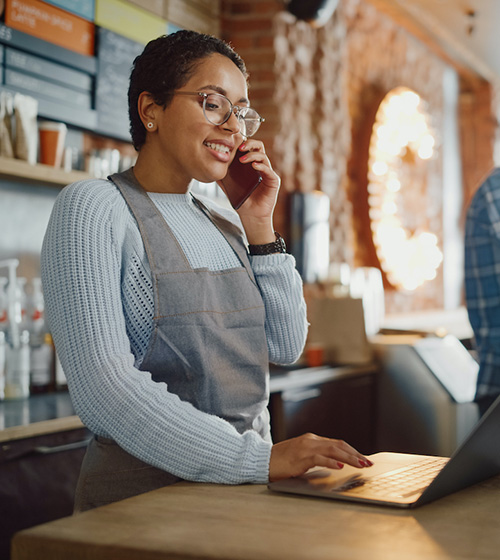 Small business owner woman wearing glasses and an apron multi tasking at a retail counter, talking on the phone and looking at a laptop.